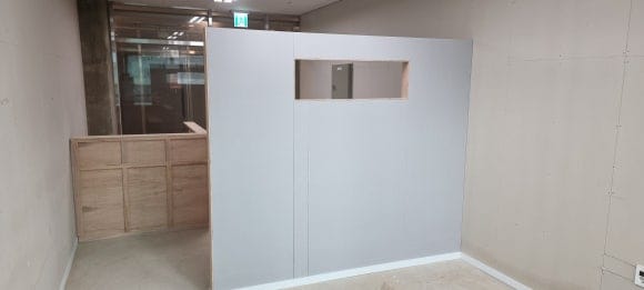 작은 사각 창을 낸 파티션 벽이 시술 공간을 가리고, 뒤쪽에 마감 전 맞춤 카운터가 함께 보이는 목공 작업 완료 전경.