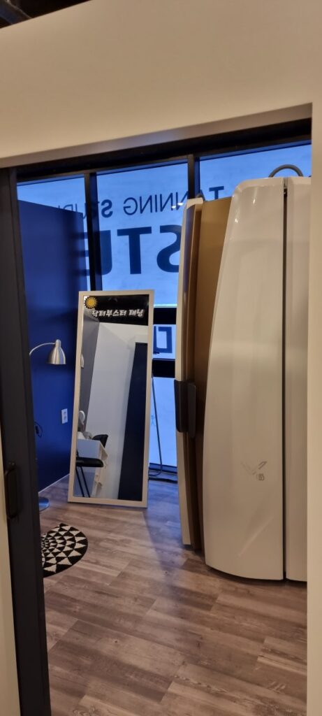 사진 14. 시술실 안에 설치된 태닝기계와 벽면 거울이 보이며, 입구에는 짙은 색 슬라이딩 도어가 열려 있습니다.