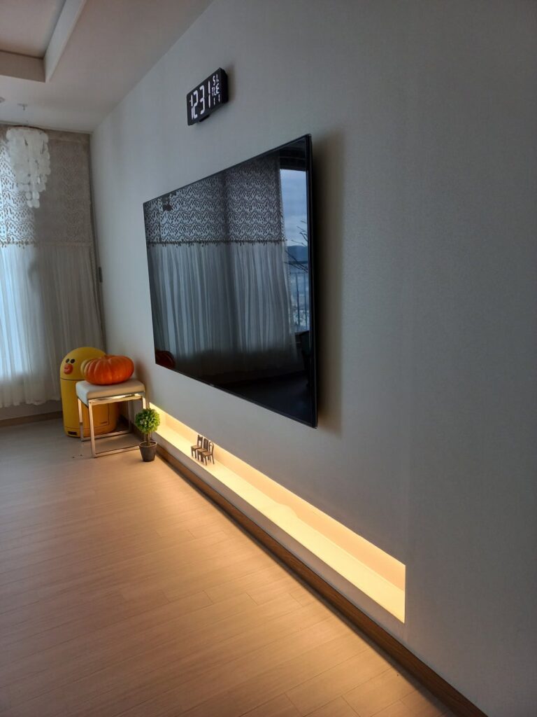 사진 3. 정면에 가까운 각도에서 본 액자형 TV 반매립 시공으로, TV가 벽면에 가깝게 붙고 아래에는 콘센트가 포함된 간접조명 선반이 이어져 있다.
