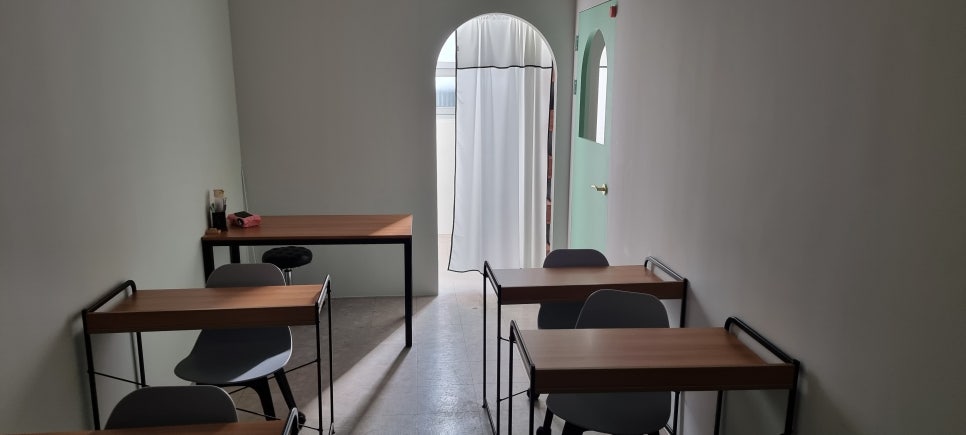 사진 14. 칠판 앞에서 원장실과 입구 방향을 바라본 강의실 내부로, 책상 사이 통로 끝에 아치형 게이트와 출입문이 이어지는 모습.