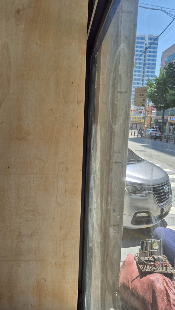 사진 23. 매장 안쪽에서 창가를 바라본 모습으로, 합판으로 마감한 벽면과 샤시 사이의 좁은 공간이 보이며 외부 도로와 차량이 함께 보입니다.